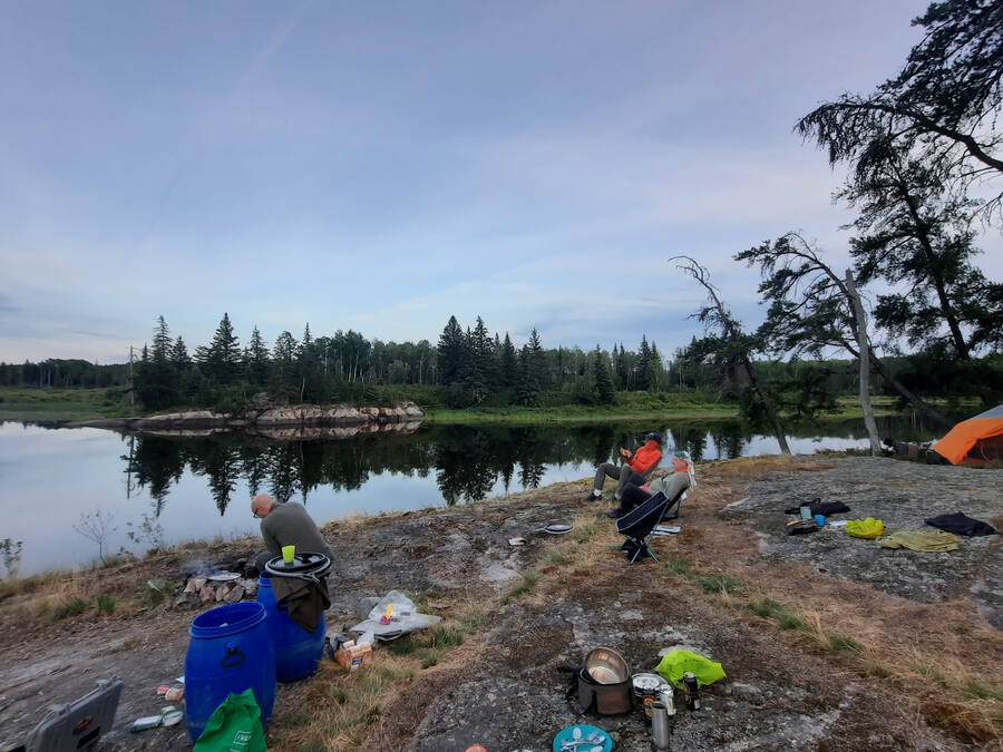 An evening camp on a granite point beside a still lake, with tents, barrels and paddlers relaxing