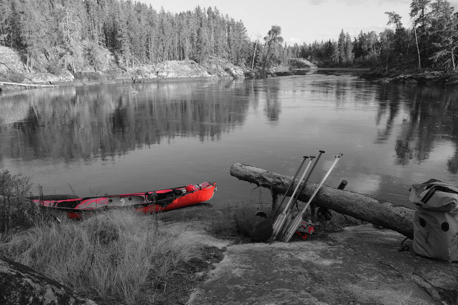 A red canoe pulled up on a granite shore beside a calm river, paddles leaning on a log