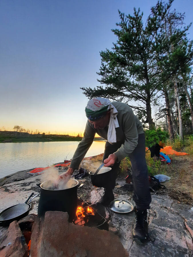 A camp cook stirring a steaming pot over a fire on a granite shore at sunset