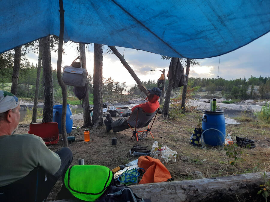 A shoreline camp under a blue tarp, with food barrels, camp chairs and gear bags